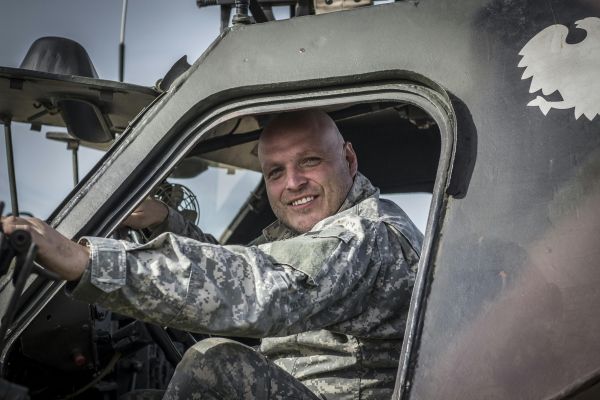 Smiling soldier in uniform inside a military vehicle cockpit on a sunny day.