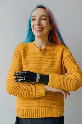 Cheerful woman with pink and blue hair wearing a prosthetic arm and yellow sweater indoors.